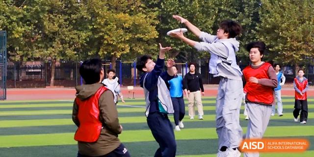 Students jump and reach for a white frisbee during a competitive outdoor sports match at Beijing Xicheng Academy.