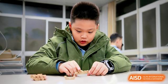A young boy in a green jacket focuses intently on building a small wooden block structure at his desk during a classroom activity at Yangjiang Yangdong Eone school.