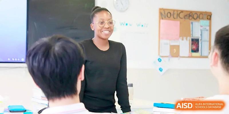 A smiling female teacher with glasses stands in a bright classroom, engaging warmly with her students during a lesson at Yangjiang Yangdong Eone school.