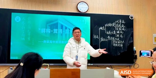 A male teacher in a white puffer jacket gives an engaging presentation in front of a chalkboard and digital display at Yangzhou Shuren High School.