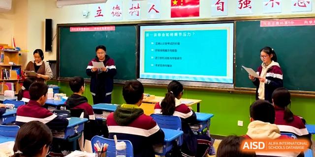 A student standing at the front of the classroom reads from a paper while a lesson outline is displayed behind her at Yangzhou Shuren High School.