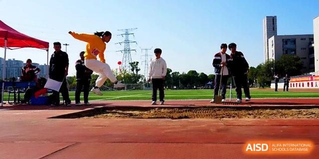 An athlete in a yellow shirt performs a long jump, soaring through the air toward the sand pit at Yangzhou Shuren High School.