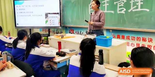 A female instructor speaks into a microphone while standing at a podium to lead a large classroom session at Yinchuan No.1 High School.