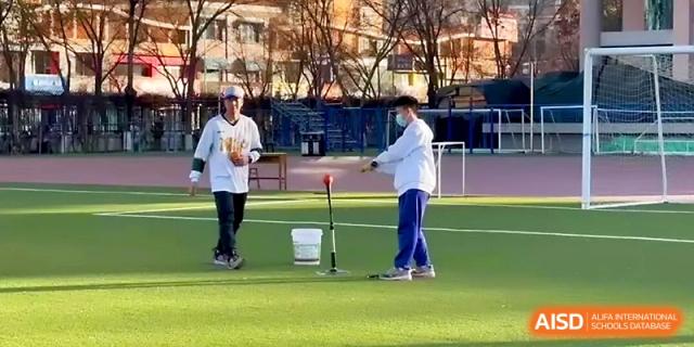 A student practices hitting a ball off a batting tee while a coach observes and provides feedback during practice at Yinchuan No.1 High School.