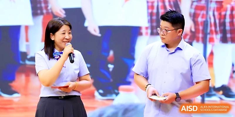 A female speaker holds a microphone while presenting alongside a male colleague on a stage at Zhangjiagang IVY Experimental School.