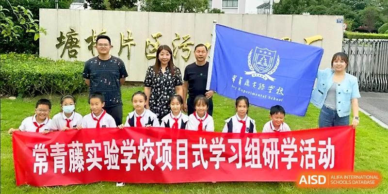 Students and faculty stand outdoors behind a red banner and a blue school flag for a group photo at Zhangjiagang IVY Experimental School.