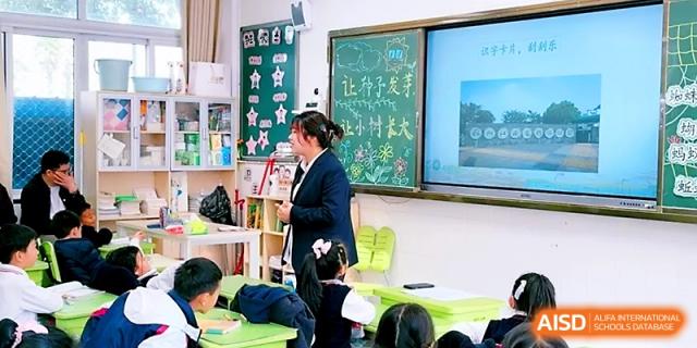 A teacher stands at the front of a primary classroom, gesturing toward a screen while students listen at Zhangjiagang IVY Experimental School.