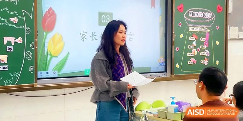 A woman stands in front of a digital screen, reading from a document during a parent-teacher meeting at Zhangjiagang IVY Experimental School.