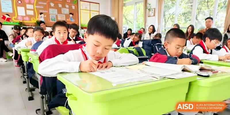 A young student in a white shirt and red scarf stays focused while writing at a bright green desk at Zhangjiagang IVY Experimental School.