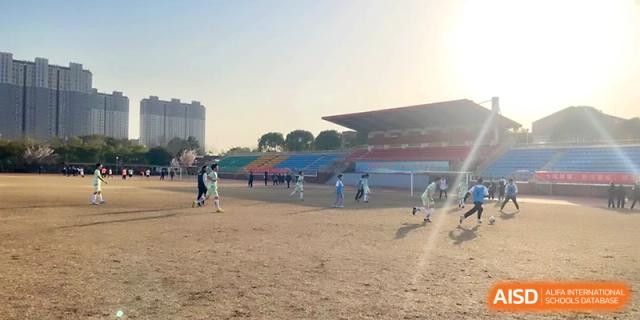 Children play a soccer match on a large dirt field with a stadium in the background at Zhangjiagang IVY Experimental School.