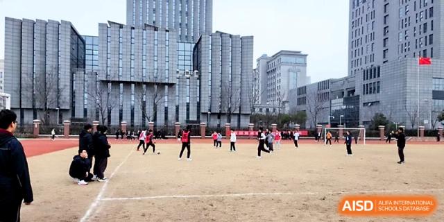 Students play soccer on a large dirt field with modern campus buildings in the background at Zhangjiagang IVY Experimental School.