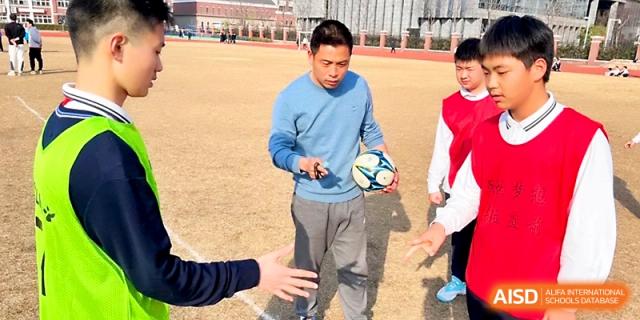 A man in a blue shirt holds a soccer ball while instructing students on the sports field at Zhangjiagang IVY Experimental School.