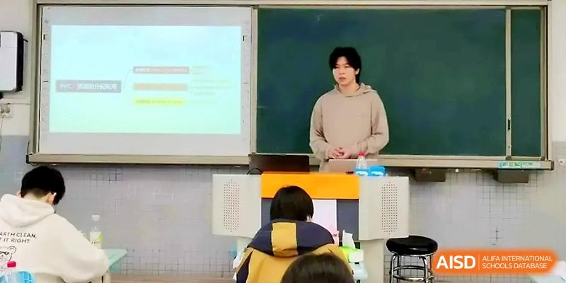 A male student stands at a podium delivering a presentation with a projector screen visible in the background at Zhengzhou No.1 High School
