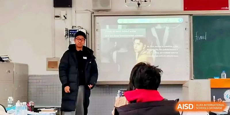 A student in a dark winter jacket stands before the class to lead a presentation or discussion in a bright room at Zhengzhou No.1 High School