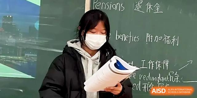 A student wearing a face mask reads from a textbook during a classroom discussion in front of a chalkboard at Zhengzhou No.1 High School