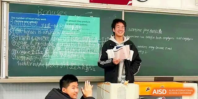 A male student smiles while holding a notebook and presenting to his classmates in a bright room at Zhengzhou No.1 High School