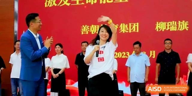 A woman waves to the crowd while speaking into a microphone on a red stage during a school event at Zhengzhou No.1 High School