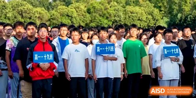 A student holds a class sign while standing in a large formation of peers on an outdoor field at Zhengzhou No.1 High School