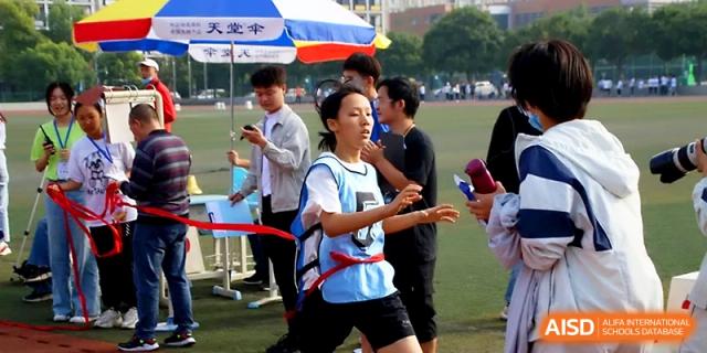 A female student in a blue vest runs during a competitive outdoor race while spectators watch from the side at Zhengzhou No.1 High School