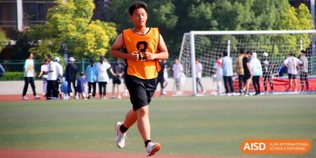 A runner in an orange jersey maintains a steady pace during a distance race on the school sports grounds at Zhengzhou No.1 High School