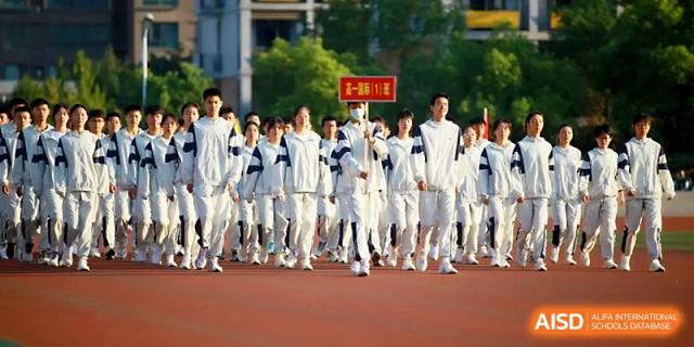 A large group of students in white tracksuits marches in formation past the stadium seating during an event at Zhengzhou No.1 High School