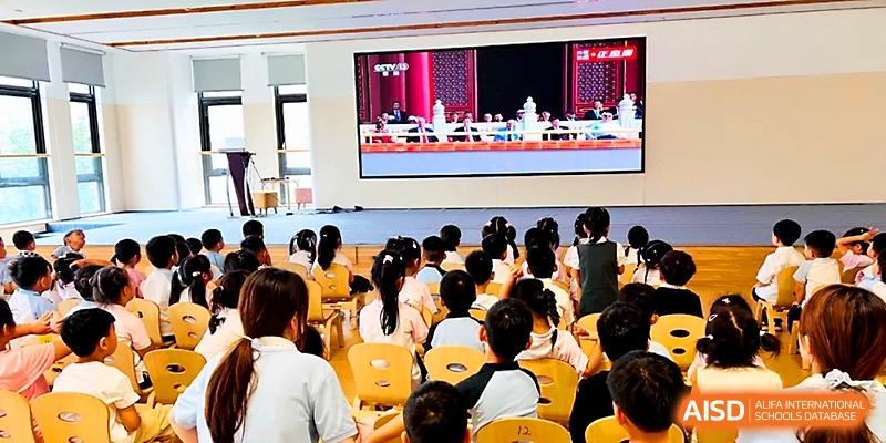 Young students sit in rows of yellow chairs watching a large screen presentation during an indoor assembly at Zhengzhou No.1&8 United International School