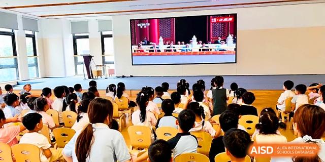 Young students sit in rows of yellow chairs watching a large screen presentation during an indoor assembly at Zhengzhou No.1&8 United International School