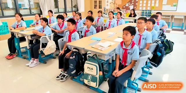 Students in light blue uniforms and red scarves sit at long wooden desks during a formal classroom session at Zhengzhou No.1&8 United International School
