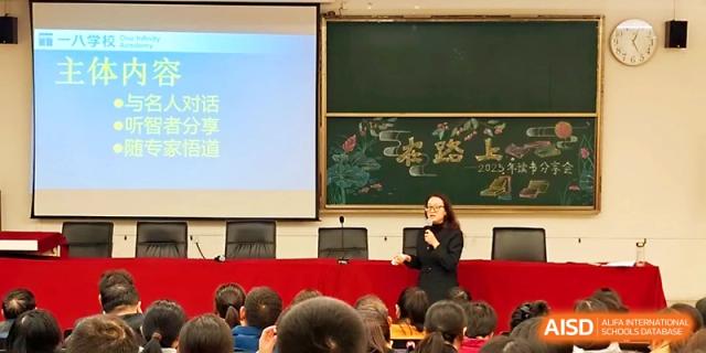 A female teacher stands at a podium delivering a lecture with a projector screen and chalkboard in the background at Zhengzhou No.1&8 United International School