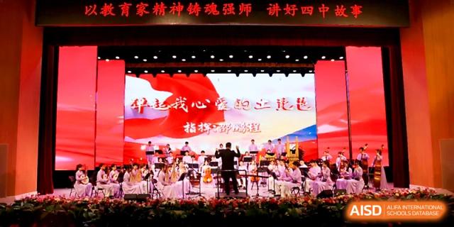 An orchestral conductor leads a large group of musicians on a decorated stage under bright lights at Zhengzhou No.4 Middle School.