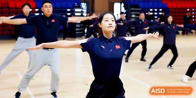 A student in a navy polo shirt performs a focused stretching or dance movement in a gymnasium at Zhengzhou No.4 Middle School.