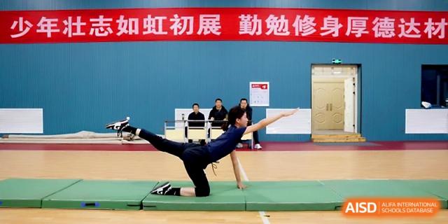 A student athlete demonstrates a balanced gymnastics pose on a green mat in a gymnasium at Zhengzhou No.4 Middle School.
