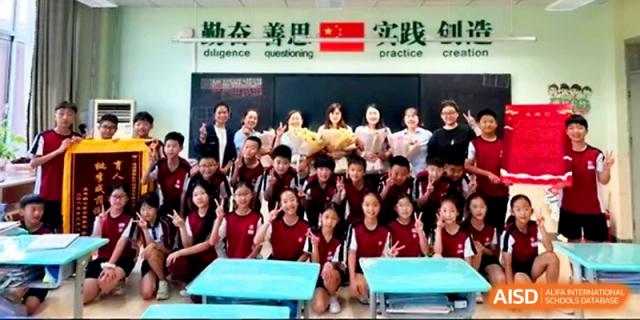 A large group of primary students and teachers pose for a class photo with flowers in a bright classroom at Zibo International Academy At Hi-Tech Zone.