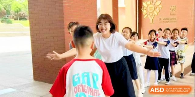 A smiling teacher in a white shirt welcomes a young student with open arms in a school hallway at Zibo International Academy At Hi-Tech Zone.