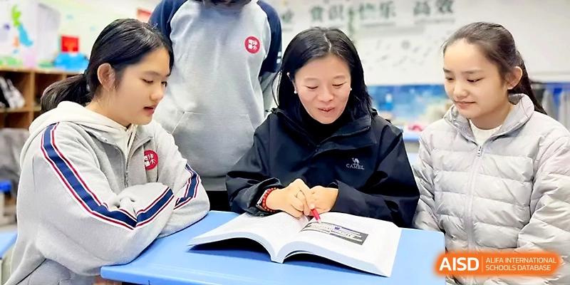 A teacher leans over a student's desk to provide personalized guidance on a textbook lesson at Zibo International Academy At Hi-Tech Zone.