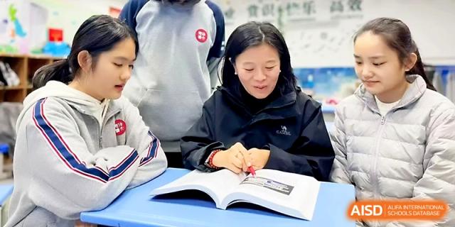 A teacher leans over a student's desk to provide personalized guidance on a textbook lesson at Zibo International Academy At Hi-Tech Zone.