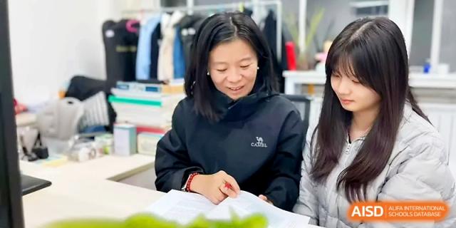 A female staff member in a black jacket smiles while reviewing educational materials in an office at Zibo International Academy At Hi-Tech Zone.