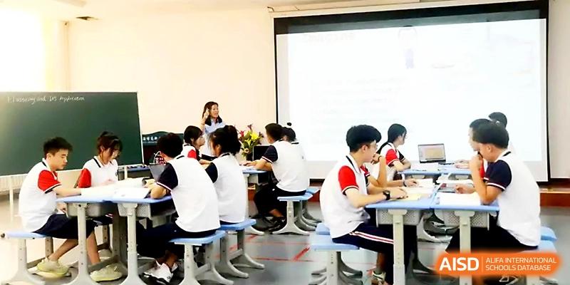 A classroom view shows students in uniforms seated at desks while a presentation is projected on a screen at Zibo International Academy At Hi-Tech Zone.