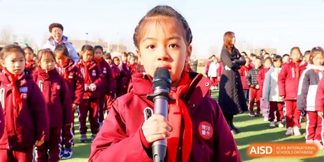 A young girl in a red uniform speaks confidently into a microphone during a school ceremony at Zibo International Academy At Hi-Tech Zone.