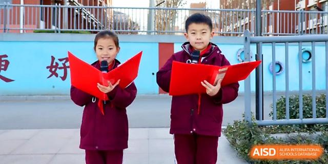 Two students hold red folders and speak into a microphone during a school presentation at Zibo International Academy At Hi-Tech Zone.