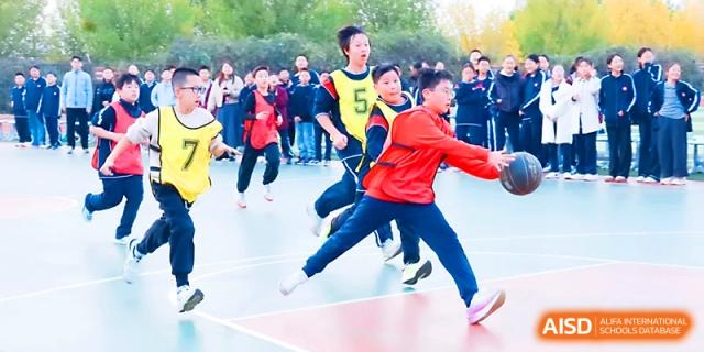 A student in a red jacket dribbles a basketball across the court during a sunny afternoon game at Zibo International Academy At Hi-Tech Zone.