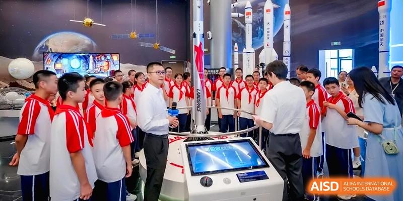 A male instructor speaks to a large group of students gathered around a space rocket model at the Affiliated High School of Fujian Normal University.