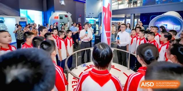A man with a microphone addresses a group of students standing in a circle around a museum exhibit at the Affiliated High School of Fujian Normal University.