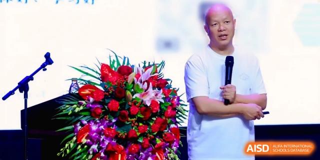 A man speaks into a microphone next to a large bouquet of flowers at a school ceremony at the Affiliated High School Of Peking University Haikou.