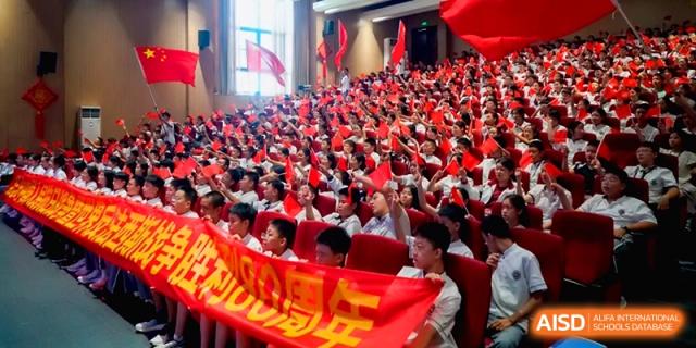 A crowded auditorium filled with students holding red banners during a patriotic event at the Affiliated High School Of Peking University Haikou.