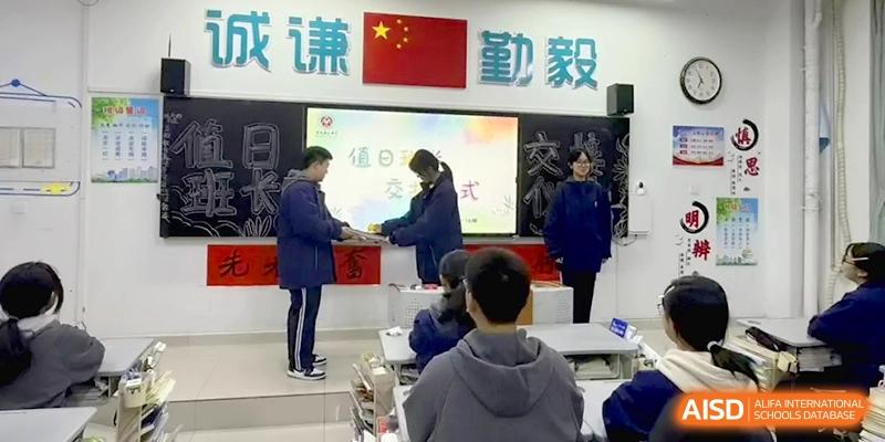 Two students stand at the front of a classroom during a formal ceremony, exchanging a book or document near the chalkboard at Baoji Nanshan High School.