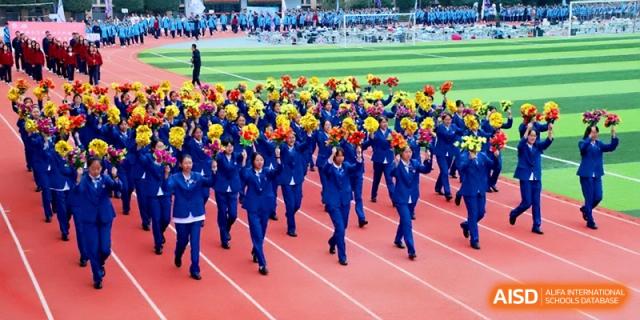 A large group of students in blue athletic wear performs a synchronized dance with yellow pom-poms on a track at Baoji Nanshan High School.
