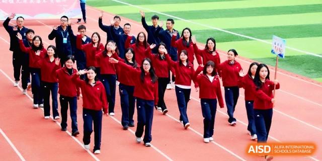 Students in red jackets wave and smile enthusiastically while marching in a formation on a green field at Baoji Nanshan High School.