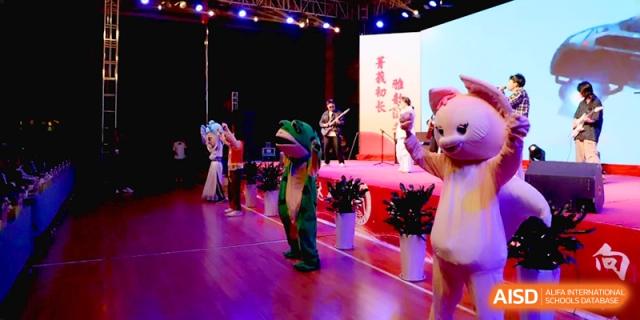Performers in full-body green and pink mascot suits put on a lively show for a seated audience in the school theater at Baoji Nanshan High School.