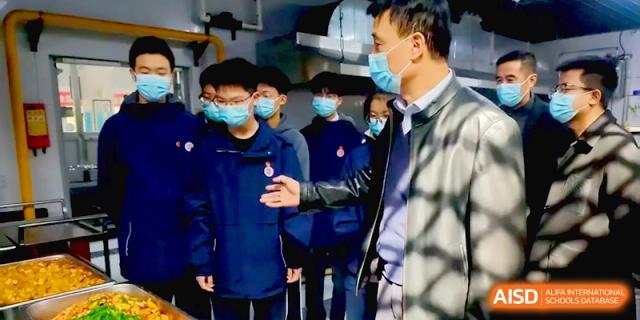 A group of students in blue jackets observes a staff member explaining kitchen safety and hygiene protocols at Baoji Nanshan High School.